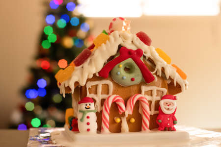 Homemade gingerbread house decorated with icing, sweets , figurines, lollies and candy canes with blurred Christmas tree on background.の写真素材