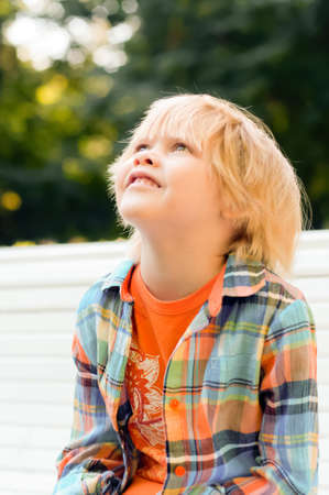 Cute blonde caucasian kid sitting on the bench looking up at the park. Summer vacation happy childhood conceptの写真素材