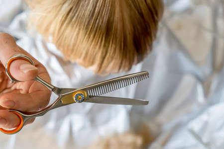 hands doing blonde toddler haircut with hairdresser scissors at home. Baby boy hair cut diy