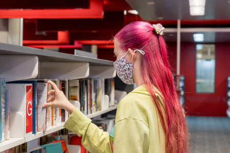 Teenage girl with ombre pink purple colored hair wearing face mask choosing books at the local...