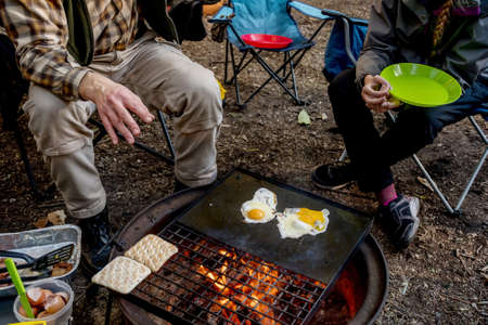 Breakfast camp cooking. Man cooking eggs and warming bread on a cast iron plate over the camp fire. Camping lifestyleの写真素材