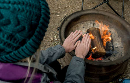 Camping lifestyle. Girl wearing beanie hat wariming hands over the campfire on a camping site. Selective focusの写真素材