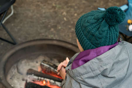 Camping lifestyle. Girl wearing beanie hat wariming hands over the campfire on a camping site. Selective focusの写真素材