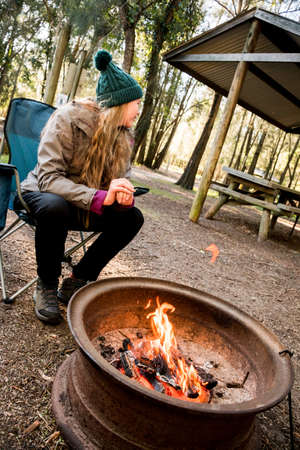Camping lifestyle . Teenage girl wearing beanie hat sitting in a camping chair near a campfire in a firepitの写真素材