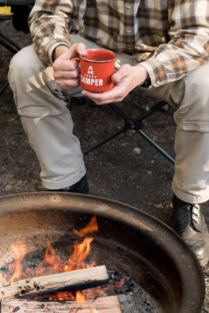 Camping lifestyle. Man wearing plaid shirt holding red enamel mug near a campfire. Happy camperの写真素材