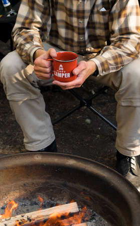 Camping lifestyle. Man wearing plaid shirt holding red enamel mug near a campfire. Happy camper