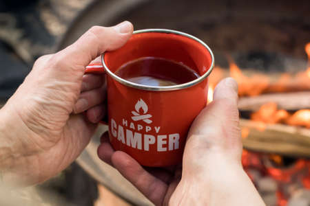 Camping lifestyle. Hands holding red enamel mug near a campfire. Happy camperの写真素材