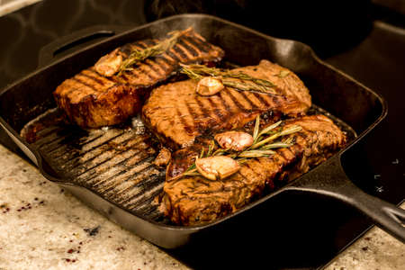 Grilling cooking New York beef sirloin strip steaks with rosemary and garlic on cast iron grill pan skillet on a ceramic stove counter top at home.の写真素材
