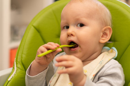 Little baby playing and feeding himself with spoon sitting in highchair. Baby first solids. Weaning. Baby boy eatingの写真素材
