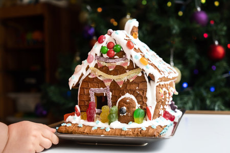Homemade gingerbread house decorated with icing, sweets, and jelly candies with blurred Christmas tree on background. Family Christmas. Toddler hand touch a gingerbread houseの写真素材