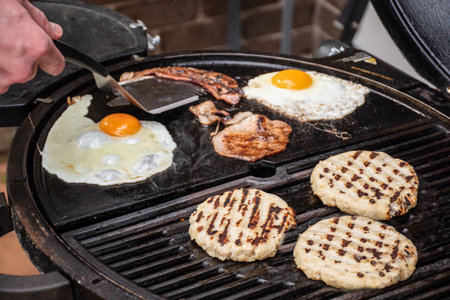 Burger patty, eggs and bacon grilling on barbecue grill plates. Backyard BBQ grill cooking burgers.の写真素材