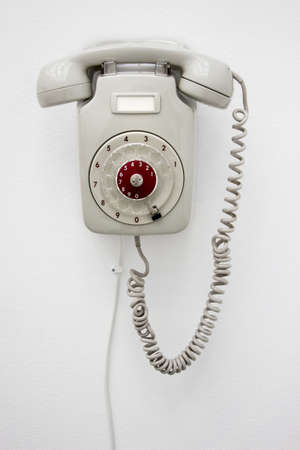 Old fashioned gray rotary telephone hanging on white wall. Vertical photo.の写真素材