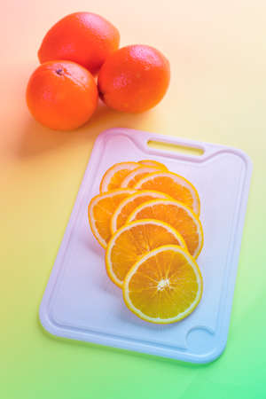 Three whole oranges with sliced oranges laying on the white cutting board on yellow background. Vibrantly colored image.の写真素材
