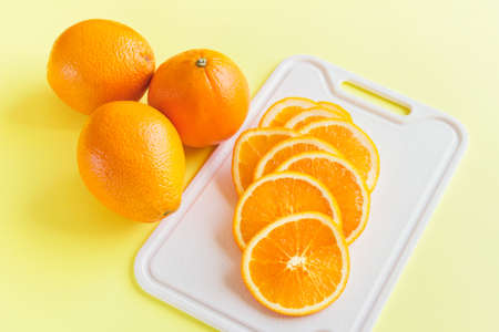 Three whole oranges with sliced oranges laying on the white cutting board on yellow background. Vibrantly colored image.の写真素材