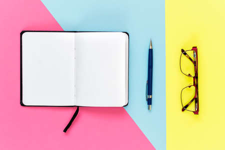 Top view of opened notebook for notes, pencil and glasses on multi colored blue, pink, yellow background.の写真素材