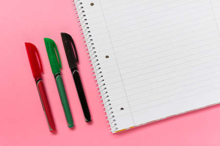 Office school supplies - three colored pens and lined spiral notebook on pink background. Photo with...