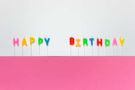 Happy Birthday greeting card with wax colorful letters candles on white pink paper background. Photo with copy blank space.の写真素材