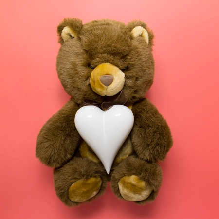 Cute furry brown teddy bear with white porcelain heart on red pink background.