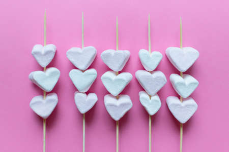 Sweet heart shaped marshmallows on wooden sticks on pink background.
