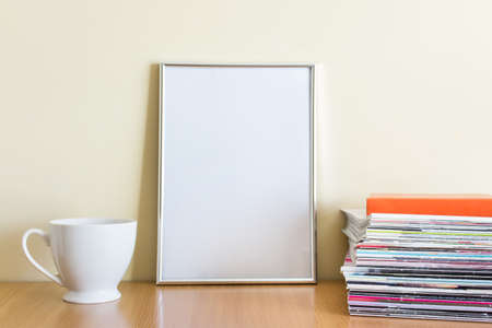 Office style composition with large A4 silver blank frame, white porcelain cup and stack of magazines on wooden surface.の写真素材