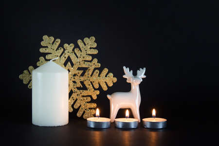 Christmas, New Year composition with three burning candles, white porcelain reindeer, golden snowflake and white large candle on black background.の写真素材
