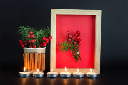 Christmas, New Year composition with many candles, frame with red background and cup with cinnamon, fir evergreen and red berries decoration on black background.の写真素材