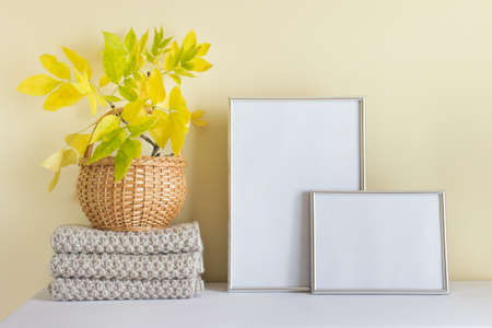 Mockup template with two silver frames vertical A4 and horizontal A5, knitted autumn warm scarf and autumn yellow leaves in wooden basket.の写真素材