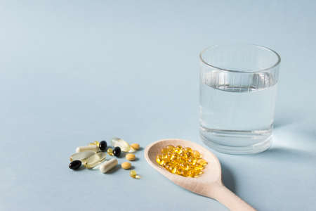 Medical composition with wooden spoon filled with vitamin capsules and glass of water on blue...