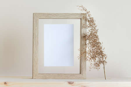 Mockup template with vertical wooden frame, white card and dry plant standing on wooden surface.