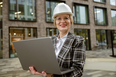 Senior female engineer in suit with white safety helmet working laptop on city building background