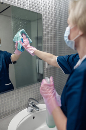Chambermaid in mask cleans mirror in hotel bathroom spraying detergent on surfaceの写真素材