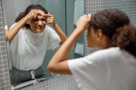 Upset young african woman removing pimple from her face while looking at mirror in bathroom homeの写真素材