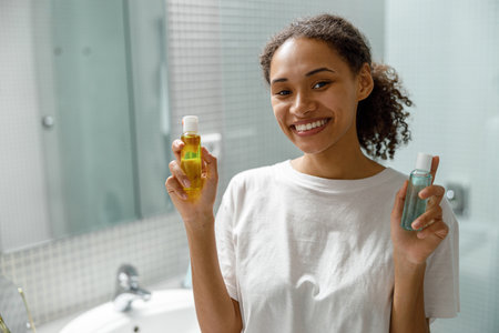 Happy african girl holding natural skincare or body care cosmetics standing in bathroomの写真素材