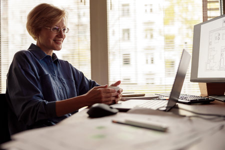 Mature smiling woman constructor drinking tea during working day from home officeの写真素材