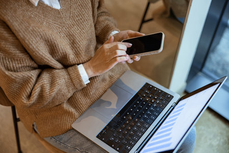 Close up of woman in casual clothes works remotely using laptop and phone while sitting in cafeの写真素材