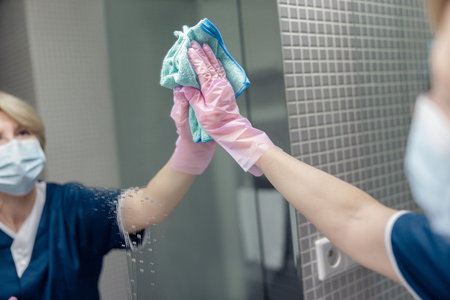 Chambermaid in mask cleans mirror in hotel bathroom spraying detergent on surfaceの写真素材