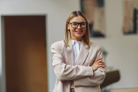Portrait of stylish smiling female enterprier standing with crossed hands on background of officeの写真素材