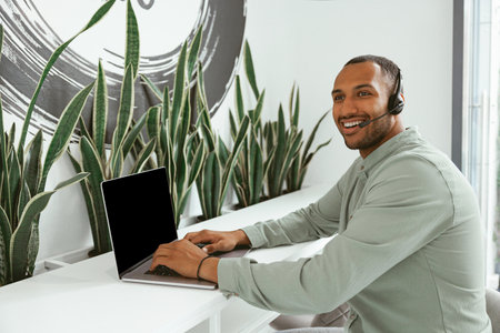 Smiling african businessman in headphones talking online with client sitting in office