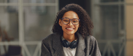 Woman freelancer with headphones smiling at camera while sitting with phone in modern coworking