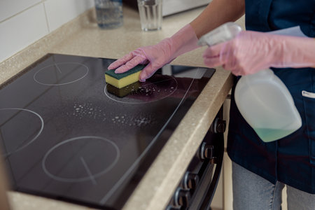 Close up of housemaid hands cleaning dirty induction stove with sponge and spray in kitchen