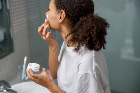 African Woman applying moisturizing cream standing behind mirror in home bathroom. Beauty procedureの写真素材