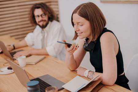 Female manager record audio message to client while sitting in coworking on colleague background