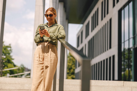 Smiling businesswoman in sunglasses is using phone while standing on modern building backgroundの写真素材