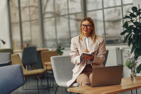 Attractive woman accountant in eyeglasses making notes during work on laptop in cozy officeの写真素材