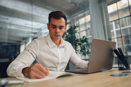 Focused male manager working on laptop and making notes while sitting in modern coworkingの写真素材