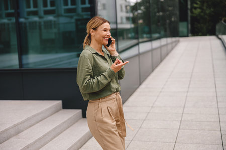 Smiling business woman is talking by phone while standing on modern building backgroundの写真素材