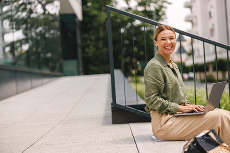 Smiling woman freelancer sitting on stairs with laptop while working on project remotelyの写真素材