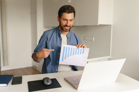 Male freelancer have video conference from laptop and showing documents while work from home kitchen