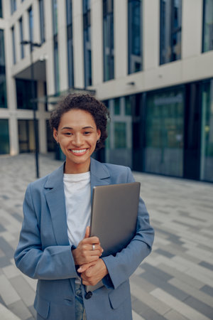 Stylish business woman with laptop standing on modern office building background and looks camera