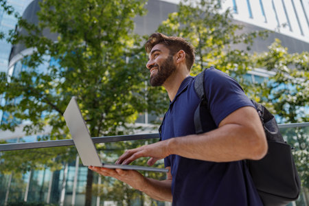 Cheerful bearded male sales manager working on laptop standing on skyscrapers backgroundの写真素材